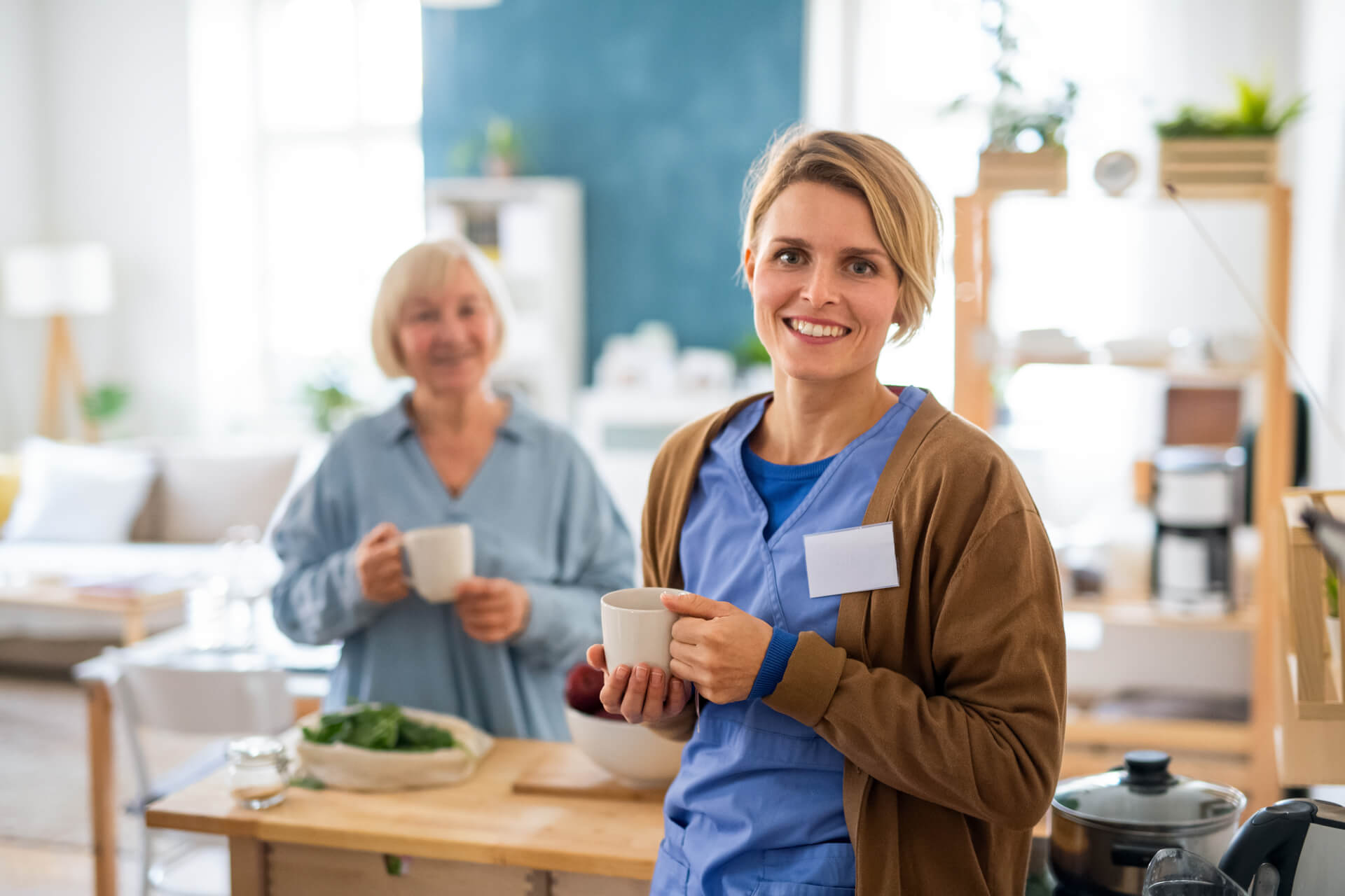 Senior woman with caregiver or healthcare worker indoors, drinking tea in kitchen.